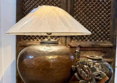 A table lamp repurposed from a unique vintage large brown ceramic pot, with antique wood carvings, in front of a pair of antique chinese shanxi door panels with lattice work